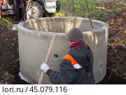 A loader lowers a concrete ring into a dug hole to build a septic tank. A worker installs a sewer into the ground. Стоковое фото, фотограф Коханова Наталья / Фотобанк Лори
