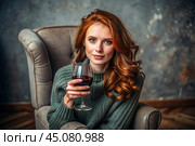 Close-up. A modern, stylish, beautiful woman with red hair and a glass of red wine in an armchair in the living room of the house. Стоковое AI-изображение, AI-художник Photojuli / Фотобанк Лори