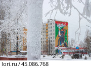 Город Тында, Амурская область, Россия. Вид на большое граффити на стене жилого дома, посвящённое строителям Байкало-Амурской магистрали (БАМ) и города Тынды. Мурал с изображением молодых энергичных людей, идущих на стройку. На заснеженной улице скульптурная композиция в виде серпа и молота. На переднем плане, покрытый инеем, ствол дерева. (2024 год). Редакционное фото, фотограф Андрей Степанов / Фотобанк Лори