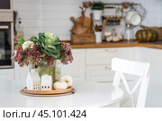 Still-life. A bouquet of decorative cabbage in a vase, pumpkins and white houses on the table in the interior of a home kitchen. A cozy autumn concept. Стоковое фото, фотограф Photojuli / Фотобанк Лори