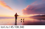 A person playing fetch with two dogs at sunset on a tranquil beach with vibrant clouds in the background. Стоковое AI-изображение, AI-художник Сергей Толмачёв / Фотобанк Лори