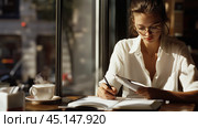 A woman in a cozy café writes in her notebook while enjoying a cup of coffee during a sunny afternoon in an urban setting. Стоковое AI-изображение, AI-художник Сергей Толмачёв / Фотобанк Лори