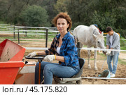 Adult female farmer working on farm tractor at stables at equestrian club. Young male worker in background cleaning up horse droppings. Стоковое фото, фотограф Яков Филимонов / Фотобанк Лори