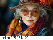 Stylish older woman in a bright and eccentric outfit. She wears a wide-brimmed leopard-print hat decorated with large flowers, as well as glasses with amber lenses, which add originality to her image. Her bright scarf with colorful patterns completes the overall ensemble. Silver hair and bright makeup emphasize her charismatic personality and confidence. The woman radiates positivity, self-expression and love for fashion. The photo conveys the spirit of creativity and vitality. Стоковое AI-изображение, AI-художник Анна Кривицкая / Фотобанк Лори
