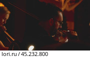 Close-up of a man playing the violin. In the background a woman with violins and cello. Warm light emphasises the violinist's instrument. A symphonic concert. Russia, September 2, 2024. Редакционное видео, видеограф Воловодов Владимир / Фотобанк Лори