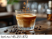The moment of pouring coffee into a glass. Coffee drops fall, creating a beautiful ripple on the surface of the drink, and in the background the interior of the cafe is blurred. Several coffee beans lie on the wooden surface of the table, which adds atmosphere and coziness. The picture conveys a feeling of freshness and vigor. Стоковое AI-изображение, AI-художник Анна Кривицкая / Фотобанк Лори