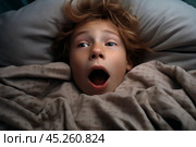 Top view, portrait of Boy with freckles lied in bed under blanket and yawns with wide opened mouth. He surprised, screaming or startled. Kid is surrounded by grey pillow and blanket. Стоковое AI-изображение, AI-художник Sergey Grishin / Фотобанк Лори