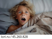 Top view, portrait of Boy with freckles lied in bed under blanket and yawns with wide opened mouth. He surprised, screaming or startled. Kid is surrounded by grey pillow and blanket. Стоковое AI-изображение, AI-художник Sergey Grishin / Фотобанк Лори