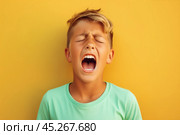 extremely whim Upset blond curly haired boy in t shirt screaming and crying or very emotional expression sings songs with wide opened mouth and closed eyes against yellow color wall. Singer audition. Стоковое AI-изображение, AI-художник Sergey Grishin / Фотобанк Лори