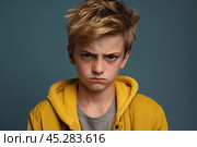 Portrait of disgruntled blond boy against grey background. The boy experiencing negative emotions due to fact that his family tore him away from computer games and forced him. Sulking Caucasian boy. Стоковое AI-изображение, AI-художник Sergey Grishin / Фотобанк Лори