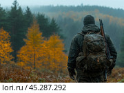 A man standing with his back to the forest in rainy weather. He is wearing camouflage clothing and a gray hat, carrying a large dirty backpack and a rifle. The autumn forest with golden and green trees creates an atmosphere of wilderness and adventure. The fog and rain give the scene an air of mystery and harshness, making the image ideal for use in survival, hunting and outdoor activities themes. Стоковое AI-изображение, AI-художник Анна Кривицкая / Фотобанк Лори