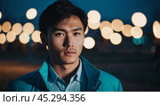 A young man with a confident expression poses against a backdrop of colorful lights at night in urban setting. A resident of Kazakhstan, a cute man of 25 years old. Student or applicant. AI generated. Стоковое AI-изображение, AI-художник Юлия Петровская / Фотобанк Лори