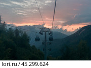 The cable car glides over lush mountains as the sun sets, painting the sky in hues of orange and pink, creating a serene, picturesque view. Стоковое фото, фотограф Алексей Климкин / Фотобанк Лори