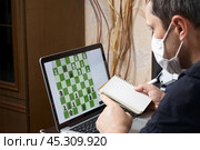 A man in a medical mask solves a chess problem online. Chess board with figures on a laptop screen. Leisure in self-isolation. International chess day. Стоковое фото, фотограф Vadzim Yakubovich / Фотобанк Лори