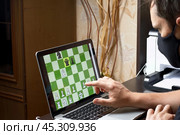 A man in a medical mask solves a chess problem online. Chess board with figures on a laptop screen. Leisure in self-isolation, in the twilight. International chess day. Стоковое фото, фотограф Vadzim Yakubovich / Фотобанк Лори