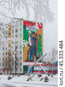 Город Тында, Амурская область, Россия. Вид на большое граффити на стене жилого дома, посвящённое строителям Байкало-Амурской магистрали (БАМ) и города Тынды. Мурал с изображением молодых энергичных людей, идущих на стройку. На заснеженной улице скульптурная композиция в виде серпа и молота. На переднем плане, покрытые инеем, ветви дерева. (2024 год). Редакционное фото, фотограф Андрей Степанов / Фотобанк Лори