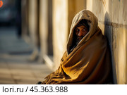 Houseless person wrapped in an old blanket sitting against a wall on a city street. concept of deprivation, vagrancy, homelessness. Стоковое AI-изображение, AI-художник 123 12 / Фотобанк Лори