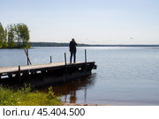 Женщина стоит на деревянном пирсе и фотографирует озеро. Турист с рюкзаком во время путешествия по сельской местности. Вид с берега на озеро. Озеро Вельё, Новгородская область, Россия. Стоковое фото, фотограф Андрей Степанов / Фотобанк Лори