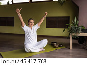 Satisfied woman practicing kundalini yoga, sitting on mat in comfortable lotus position with arms raised up doing ego or personality mudra, exercising in white clothes in room. Стоковое фото, фотограф Сергей Чайко / Фотобанк Лори