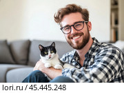 Young handsome smiling brunette man wearing glasses and checkered shirt bonding his tabby cat in hands on room background. Good friends. Friendship of pet and its owner. Cuddles. Copy space for text. Стоковое AI-изображение, AI-художник Марина Демидюк / Фотобанк Лори