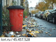 A city street with trash scattered around a red trash container. The container is on the sidewalk, next to bushes, and parked cars are visible in the distance. The street looks neglected, probably due to insufficient cleaning or a lack of waste management culture. The atmosphere is cloudy, conveying an autumn mood with fallen leaves and a gray sky. The photo symbolizes the problem of urban pollution and the need to increase environmental awareness. Стоковое AI-изображение, AI-художник Анна Кривицкая / Фотобанк Лори