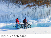 BAIKAL LAKE, KHARANTSY VILLAGE, IRKUTSK REGION, RUSSIA - February 23, 2023: Couple of tourists on bicycles travel along Small Sea and take pictures of beautiful icy rocks of Olkhon Island. Редакционное фото, фотограф Виктория Катьянова / Фотобанк Лори