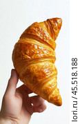 Hand holding a freshly baked croissant with golden layers in a minimalistic setting. Стоковое AI-изображение, AI-художник Юлия Гапеенко / Фотобанк Лори