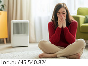The woman sneezes her face in her hands at home. Sick young woman blowing her nose. Female sneezing sitting on the floor with heater. Allergy, cold or flu. Hay fever, viral infection, health problem. Стоковое AI-изображение, AI-художник Марина Демидюк / Фотобанк Лори