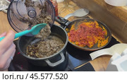 A woman transfers chopped mushrooms from a blender bowl to a saucepan with fried onions. Nearby, in a frying pan, is fried carrot with tomato paste. She is preparing mushroom caviar. Стоковое видео, видеограф Vadzim Yakubovich / Фотобанк Лори