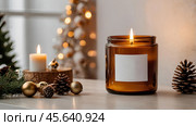 A mock-up of a cozy burning candle made of amber glass with an empty white label in a festive, New Year's home interior. Стоковое AI-изображение, AI-художник Photojuli / Фотобанк Лори