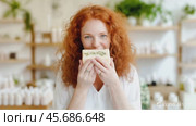 Smiling red-haired woman holding and smelling a handmade soap bar in a bright natural cosmetics shop, surrounded by eco-friendly products. Стоковое AI-видео, аниматор Башкирова Анна / Фотобанк Лори
