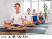 Middle-aged woman, man, preteen girl and boy practicing Padmasana with crossed legs on mats during family Hatha yoga class in wellness studio. Стоковое фото, фотограф Яков Филимонов / Фотобанк Лори