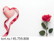 Красная роза и сердце с лентой на светлом фоне. Красная роза с зелёными листьями, расположенными справа, а слева — розовое сердце, обвязанное атласной красной лентой. Открытка ко дню святого валентина. Стоковое AI-изображение, AI-художник Анна Чижова / Фотобанк Лори