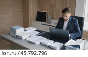 Businessman working on laptop at office desk with piles of paperwork. Стоковое видео, видеограф Данил Руденко / Фотобанк Лори