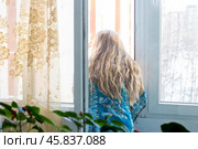 Woman with long blonde hair stands near an open window and looks out into the street, home quarantine self-isolation. Стоковое фото, фотограф Коханова Наталья / Фотобанк Лори