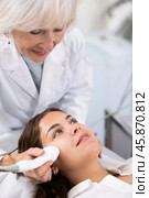 Professional mature female cosmetologist doing facial skin ultrasound cavitation for flawless woman face laying in beauty salon. Стоковое фото, фотограф Яков Филимонов / Фотобанк Лори
