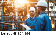Engineers in blue overalls,  white helmet and glasses, holding a clipboard talking at the construction site, illuminated by sunlight. Shallow depth of field. Стоковое AI-изображение, AI-художник Михаил Строгалев / Фотобанк Лори