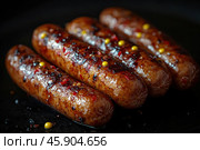 Close-up of delicious fried sausages on a dark background. Their surface is shiny with oil and covered with a golden crust with seasonings such as mustard, black pepper and pieces of red pepper. The sausages look juicy and soaked in spices, which emphasizes their piquant and rich taste. Detail allows you to see the texture of the casing and spicy accents, which makes the image especially appetizing. Стоковое AI-изображение, AI-художник Анна Кривицкая / Фотобанк Лори