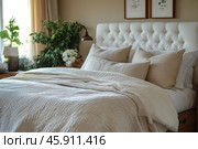 A cozy and bright bedroom. The central place is occupied by a large bed with a soft cream-colored headboard, made using the capitonné technique (button quilting). On the bed lies a light quilted bedspread, neatly folded, and several decorative pillows in light beige tones, which create comfort and a feeling of warmth. To the left of the bed, you can see a wooden bedside table with decorative elements such as a vase with a plant. In the background, there are indoor plants that enliven the interior and add green accents. The walls are decorated with minimalist paintings with plant motifs. Natural light floods the room through the window, giving it freshness and tranquility. Стоковое AI-изображение, AI-художник Анна Кривицкая / Фотобанк Лори