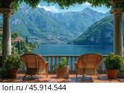 A picturesque terrace overlooking a tranquil lake surrounded by green mountains. In the foreground are two wicker rattan chairs standing on a terracotta tiled floor. Between the chairs is a wicker pot with a green plant, and on either side of the terrace are two pots with colorful flowering plants. A stone balustrade encloses the terrace, opening up a panoramic view of the blue surface of the lake and picturesque hills with houses on the far shore.The mountains are covered with dense vegetation, their peaks and slopes create a sense of grandeur and tranquility. The sky is clear, blue, with light clouds, illuminating the entire scene with soft sunlight. Vines hanging from the roof frame the view, adding coziness and natural harmony. Стоковое AI-изображение, AI-художник Анна Кривицкая / Фотобанк Лори