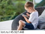 A 5-year-old little boy with a smartphone in his hands on the veranda. The child is playing on the phone. Harm of gadgets for children. Protect your child from the smartphone. Kids and gadgets. Стоковое фото, фотограф Photojuli / Фотобанк Лори
