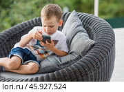 A 5-year-old little boy with a smartphone in his hands on the veranda. The child is playing on the phone. Harm of gadgets for children. Protect your child from the smartphone. Kids and gadgets. Стоковое фото, фотограф Photojuli / Фотобанк Лори