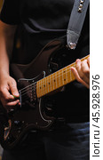 Close-up detail of black electric guitar with maple neck being played with green pick, featuring studded gray guitar strap, single-coil pickups, and chrome hardware against dark clothing background. Стоковое фото, фотограф Воловодов Владимир / Фотобанк Лори