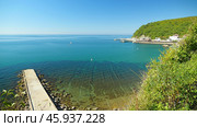 A panoramic view of a picturesque coastline with clear turquoise waters. A concrete pier extends into the sea, and a small town with buildings is nestled on the hillside. Стоковое фото, фотограф Воловодов Владимир / Фотобанк Лори