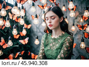 Portrait of a beautiful girl in a green dress among many suspended light bulbs and butterflies fluttering around her. Daydreaming. A magical atmosphere. Стоковое AI-изображение, AI-художник Photojuli / Фотобанк Лори