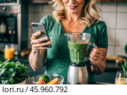 An elderly woman in the kitchen is looking at healthy food recipes on her phone and preparing a vegetable smoothie made from broccoli in a blender. Healthy detox diet drink. Стоковое AI-изображение, AI-художник Photojuli / Фотобанк Лори