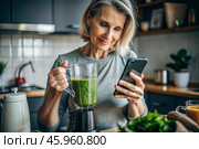 An elderly woman in the kitchen is looking at healthy food recipes on her phone and preparing a vegetable smoothie made from broccoli in a blender. Healthy detox diet drink. Стоковое AI-изображение, AI-художник Photojuli / Фотобанк Лори