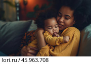 A young biracial mother lovingly holds her sleeping baby close in their cozy living room during a tranquil evening. Стоковое AI-изображение, AI-художник Sergey Grishin / Фотобанк Лори