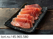Thin slices of coppa on a black rectangular plate resting on a dark wooden table. Стоковое AI-изображение, AI-художник Иван Рябоконь / Фотобанк Лори