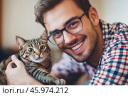 Young handsome smiling brunette man wearing glasses and checkered shirt bonding his tabby cat in hands on room background. Good friends. Friendship of pet and its owner. Cuddles. Copy space for text. Стоковое AI-изображение, AI-художник Марина Демидюк / Фотобанк Лори